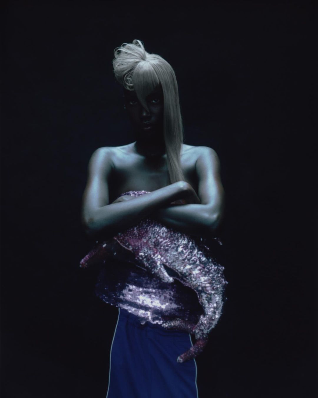 Woman with silver body paint and sculptural blonde hair poses with arms crossed against a dark backdrop in studio setting.