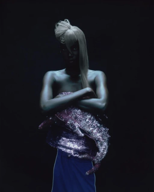 Woman with silver body paint and sculptural blonde hair poses with arms crossed against a dark backdrop in studio setting.