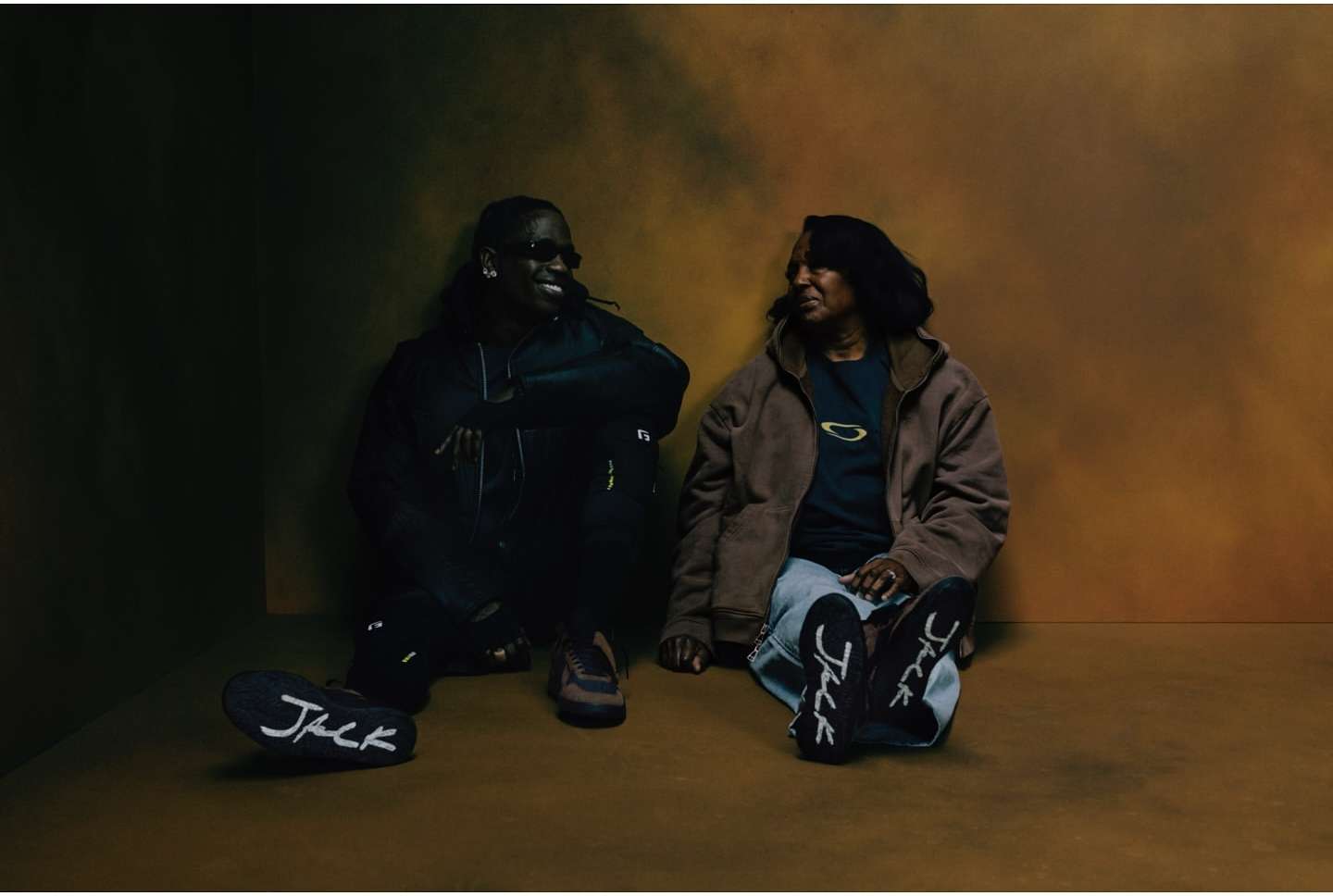 Travis Scott and an older woman seated on a studio floor against a warm mottled brown backdrop, photographed by Gabriel Moses.