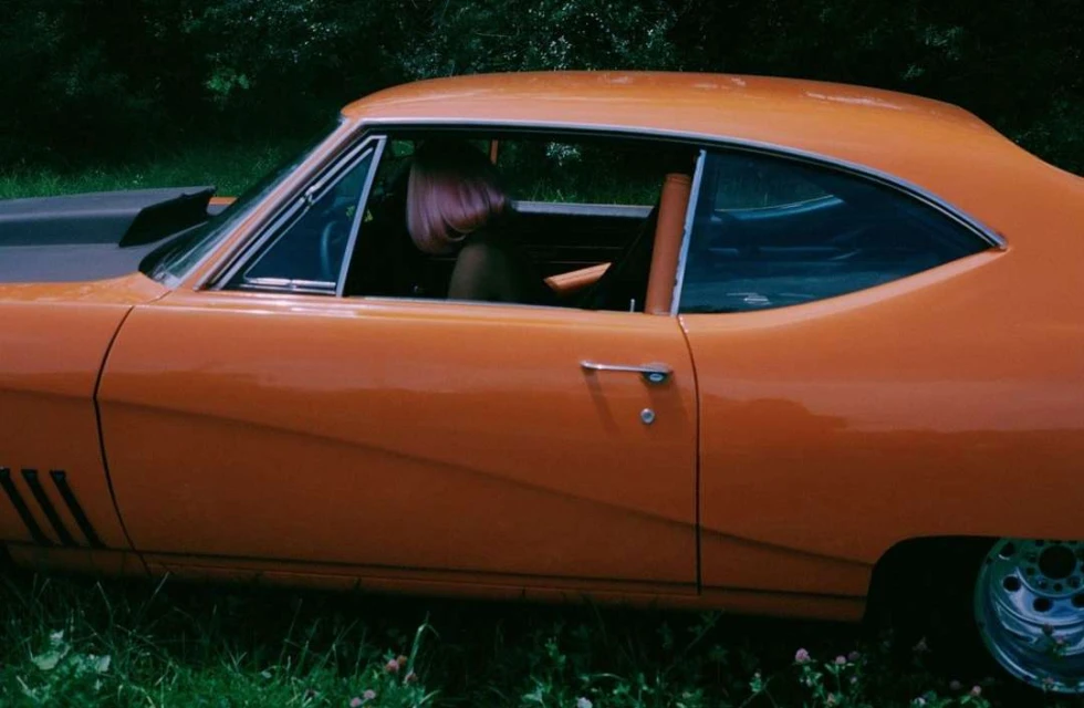 Vintage orange muscle car parked outdoors with a glimpse of a person with pink hair behind the wheel, set against a backdrop of green foliage.