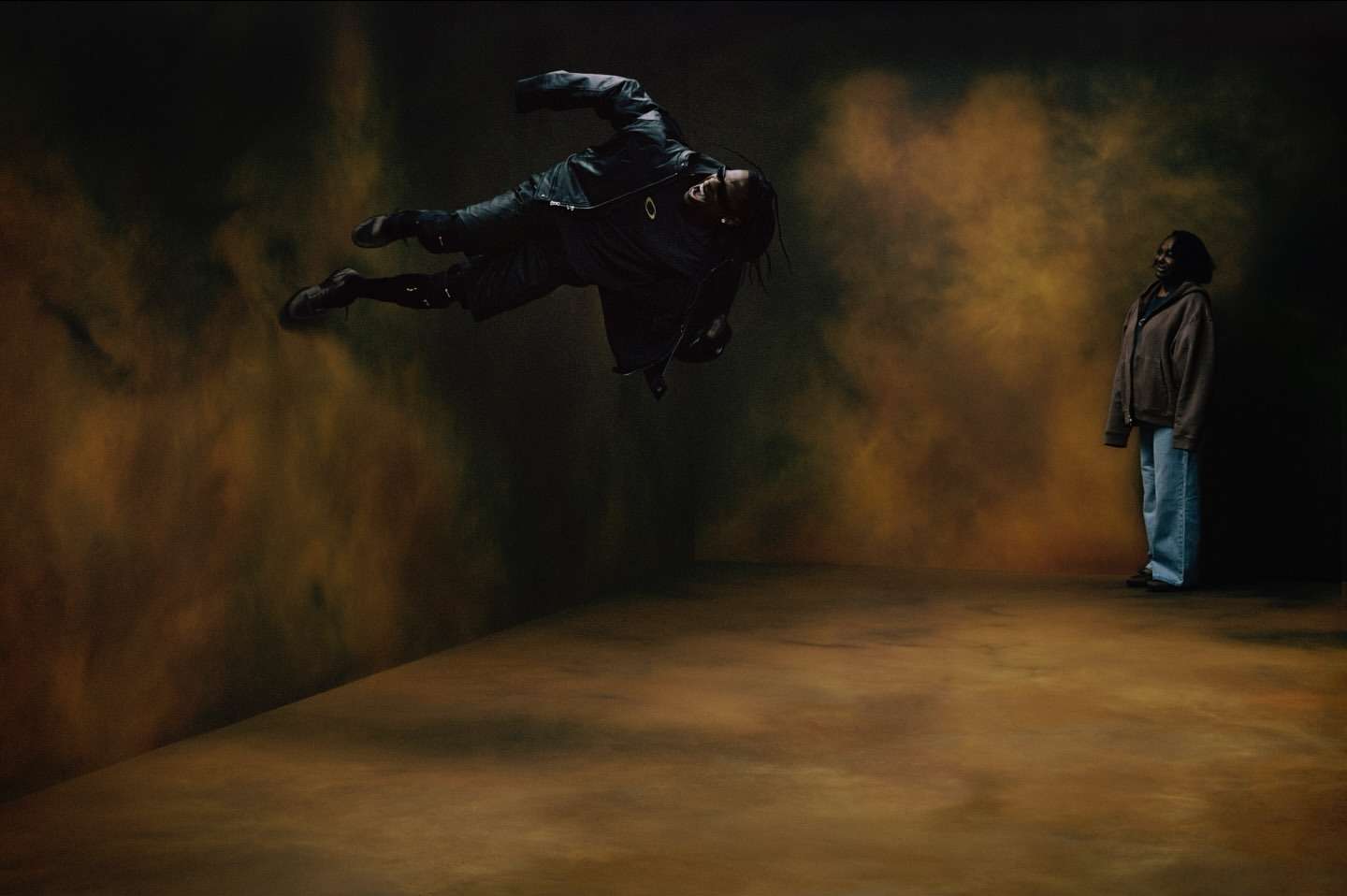 Adult male floating mid-air in a black jacket, with a second adult female in a brown hoodie and blue jeans in a studio setting.