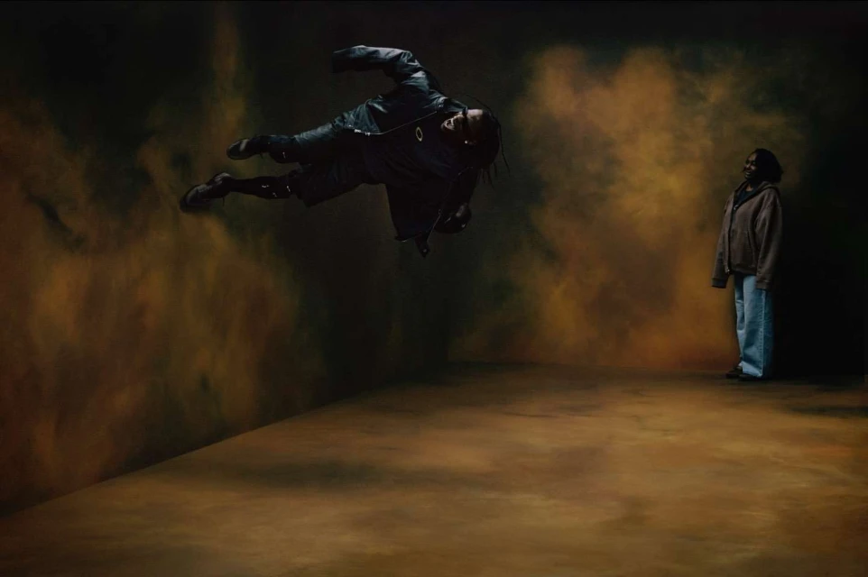 Adult male floating mid-air in a black jacket, with a second adult female in a brown hoodie and blue jeans in a studio setting.