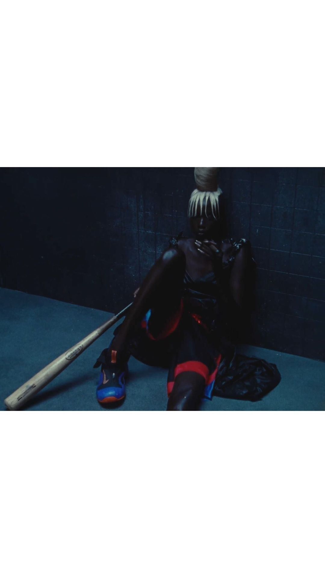 Unique fashion portrait of an adult female sitting with a baseball bat wearing athletic shoes, shot indoors against a dark wall.