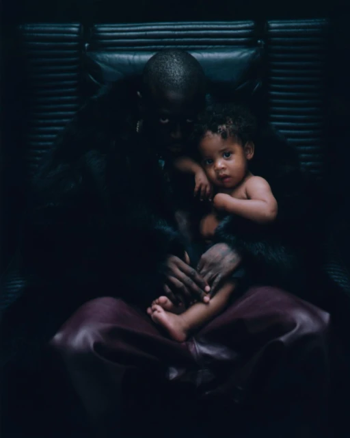 Photo of an adult male holding a toddler with a dark indoor background. The subjects sit in a black leather chair. Dark lighting is present.