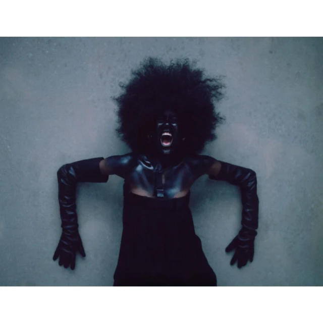 Dramatic portrait of an adult woman with afro, body paint, and gloves, posed against a gray background with an open mouth expression.