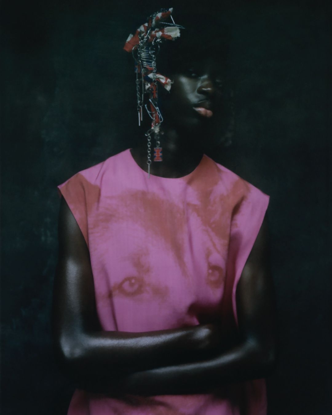 Portrait of a dark-skinned person with crossed arms, wearing a pink shirt with a face print, adorned with a headpiece against a dark backdrop.