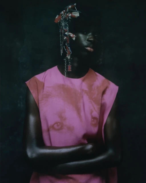 Portrait of a dark-skinned person with crossed arms, wearing a pink shirt with a face print, adorned with a headpiece against a dark backdrop.