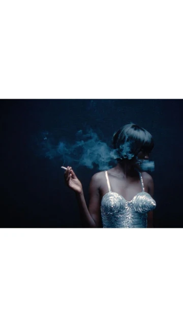 Woman in silver corset smoking a cigarette against a dark background with a blue haze of smoke around her face and short dark hair.