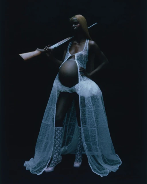 Pregnant woman with blonde hair poses with a rifle in a sheer white dress and patterned boots against a dark background in a studio.