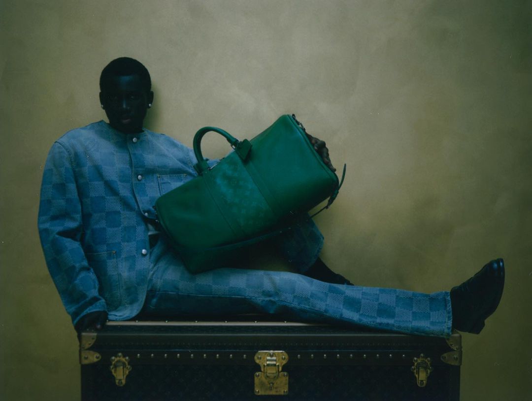 A Black male model sits on a steamer trunk holding a green leather bag, wearing a blue checker-pattern jacket and pants against a gold backdrop.