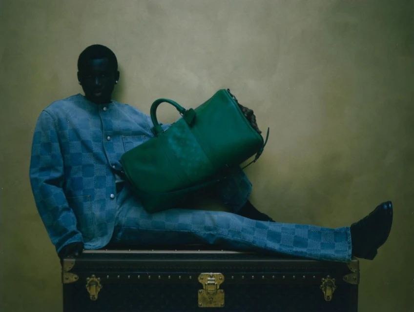 A Black male model sits on a steamer trunk holding a green leather bag, wearing a blue checker-pattern jacket and pants against a gold backdrop.