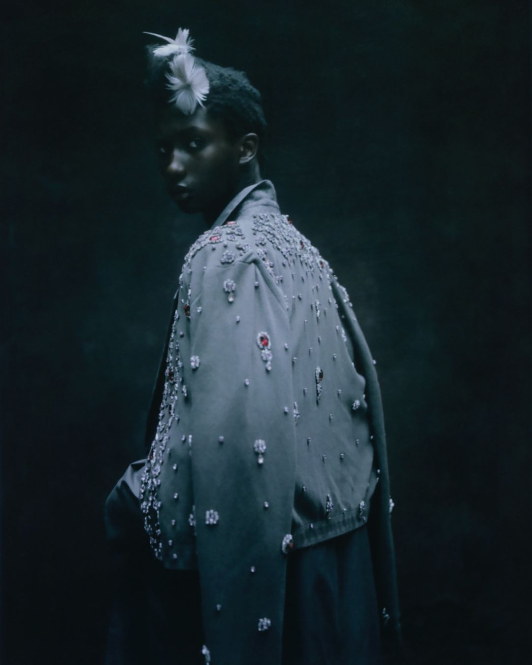 Fashion shot of a Black model wearing a beaded jacket and feathered headpiece looking over their shoulder in a dark indoor studio setting.
