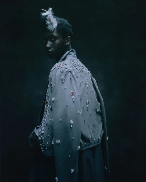 Fashion shot of a Black model wearing a beaded jacket and feathered headpiece looking over their shoulder in a dark indoor studio setting.
