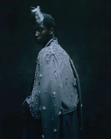 Fashion shot of a Black model wearing a beaded jacket and feathered headpiece looking over their shoulder in a dark indoor studio setting.