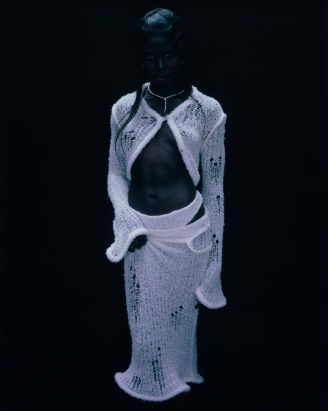 Woman with dark skin poses in a white knitted outfit against a dark background, showcasing a unique fashion style in a studio setting.