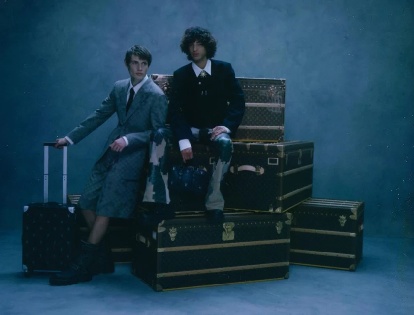 Two men in stylish suits sit on black luggage in a studio setting, posing for a fashion-forward shot with cool tones and textured backgrounds.