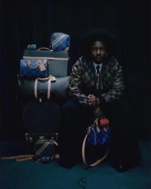 Man with afro hairstyle sits next to a stack of luxury monogrammed luggage in a dark, moody indoor setting for a fashion editorial.