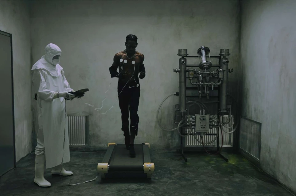 Athletic Black man runs on a treadmill wearing fitness trackers; a scientist in a hazmat suit observes in an indoor clinical setting.