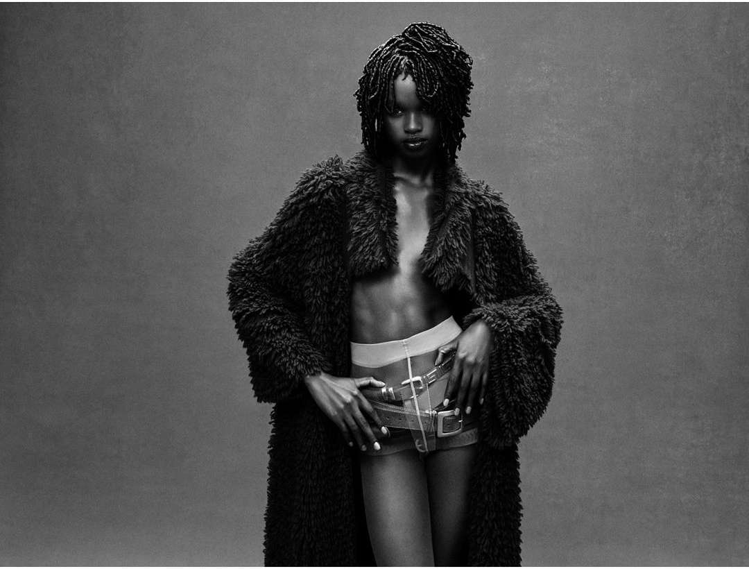 Black and white portrait of a Black woman in a faux fur coat, posing with hands on her hips. She's wearing shorts and light-colored underwear.