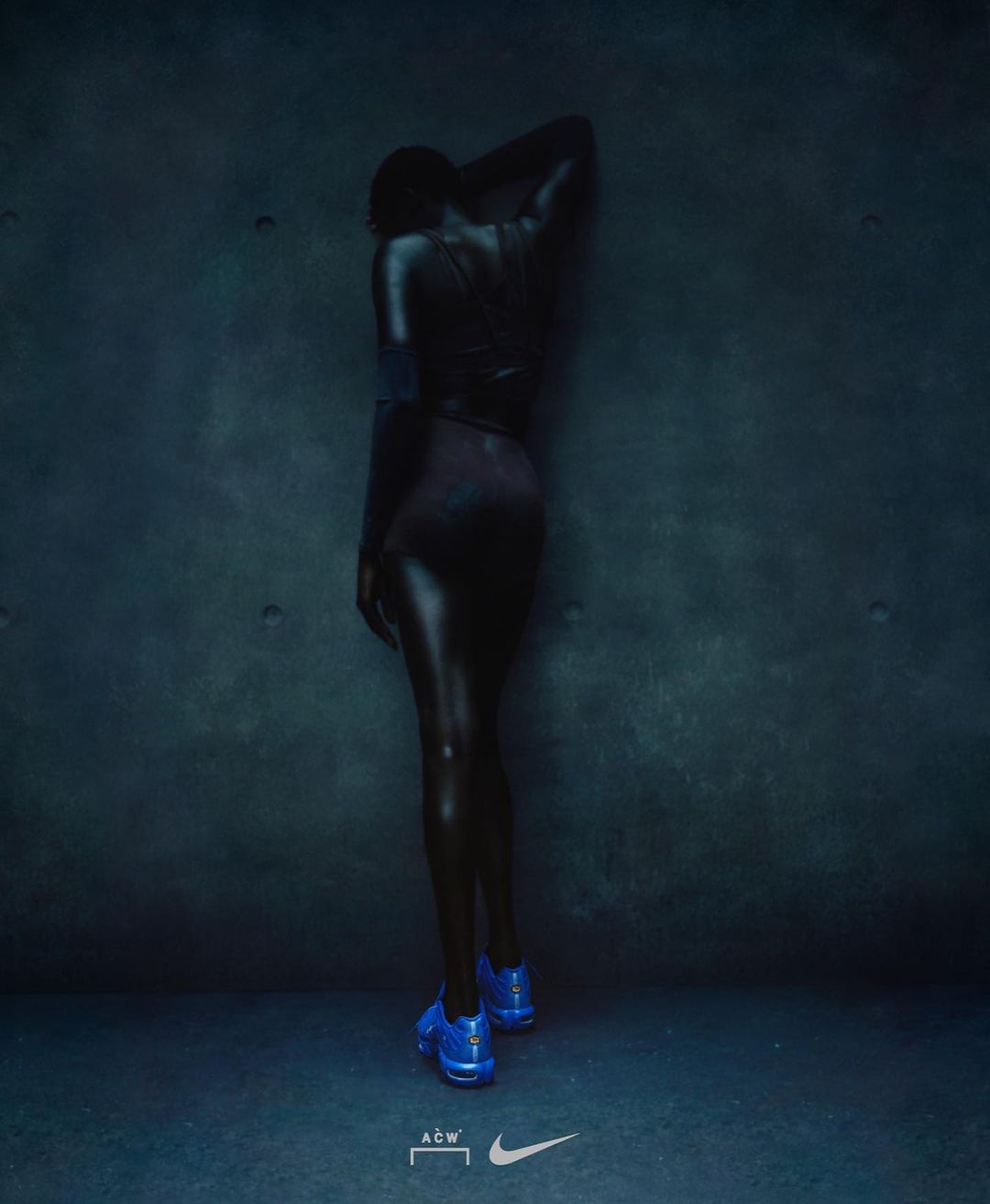 Back view of a person with dark skin and blue shoes posing against a teal textured wall in dim lighting; athletic fashion.