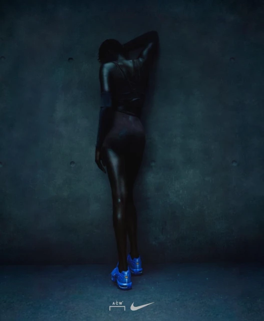 Back view of a person with dark skin and blue shoes posing against a teal textured wall in dim lighting; athletic fashion.