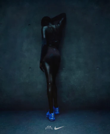 Back view of a person with dark skin and blue shoes posing against a teal textured wall in dim lighting; athletic fashion.