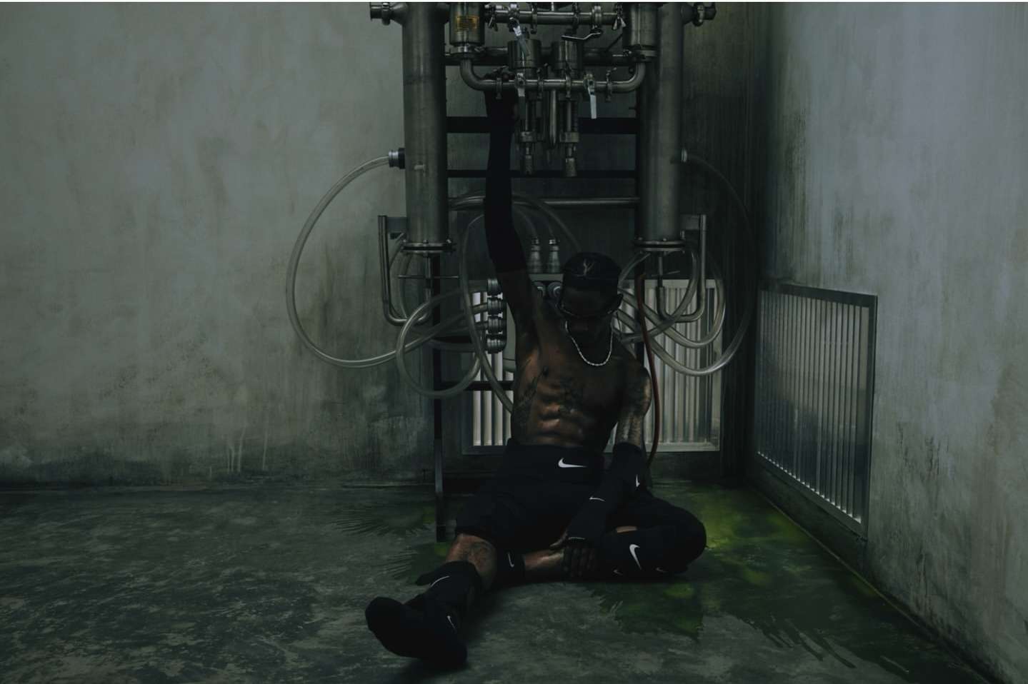 Athletic man in black sportswear poses near industrial metal pipes in an urban setting with concrete walls and green-tinted flooring.