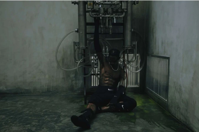 Athletic man in black sportswear poses near industrial metal pipes in an urban setting with concrete walls and green-tinted flooring.