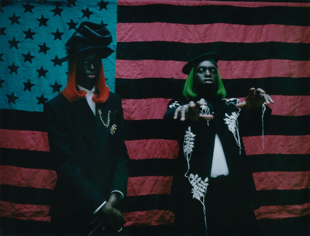 Two models wearing black suits and hats pose in front of an American flag with black stripes and blue star field.