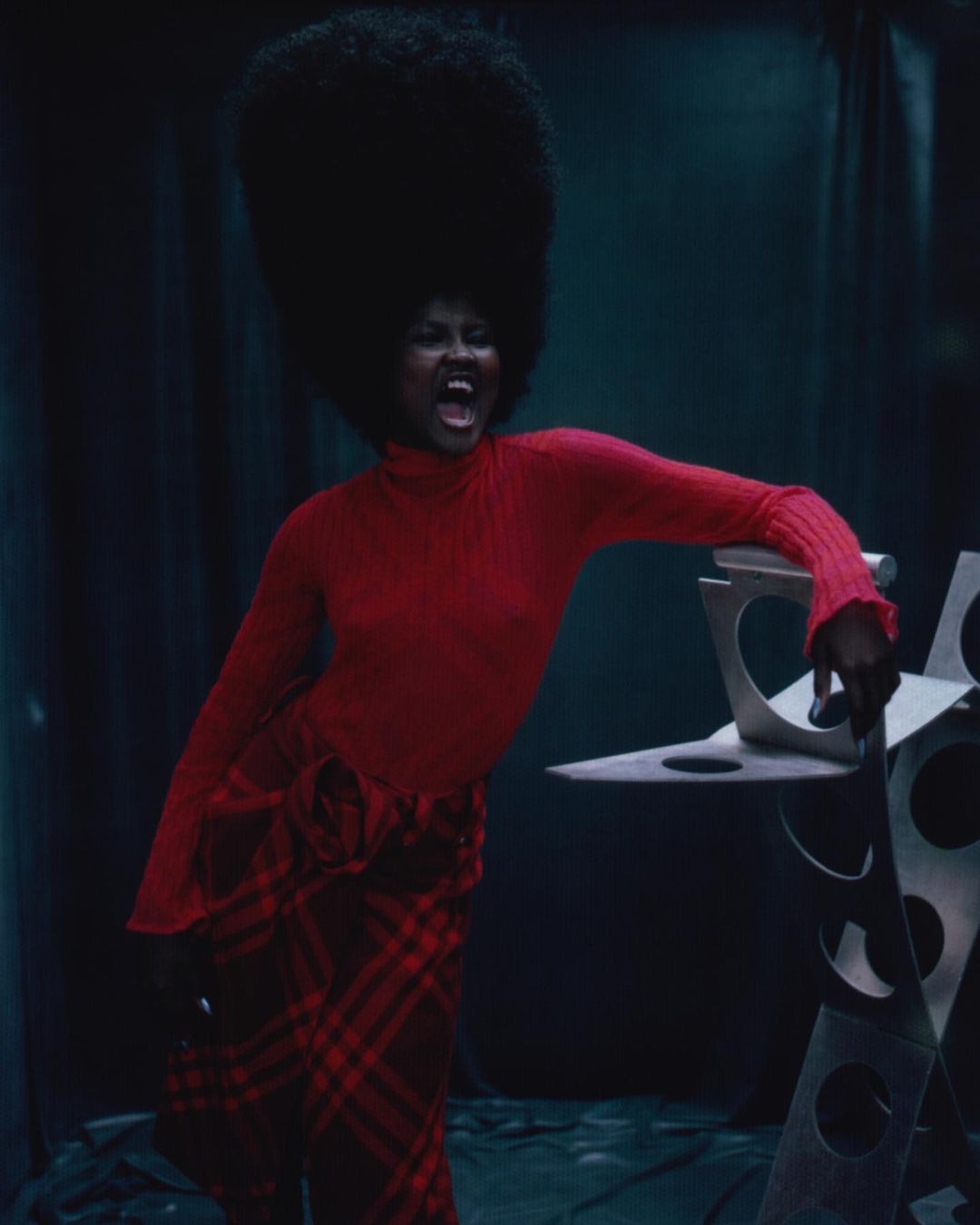 Fashion photo of Black woman with large afro wearing a red turtleneck and plaid skirt, leaning on a geometric silver sculpture.