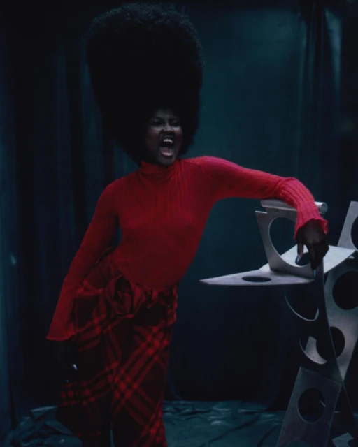 Fashion photo of Black woman with large afro wearing a red turtleneck and plaid skirt, leaning on a geometric silver sculpture.
