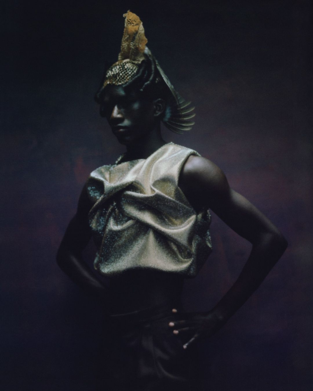 Person in a decorative golden headdress and draped metallic top poses against a dark purple backdrop in a high-fashion portrait.