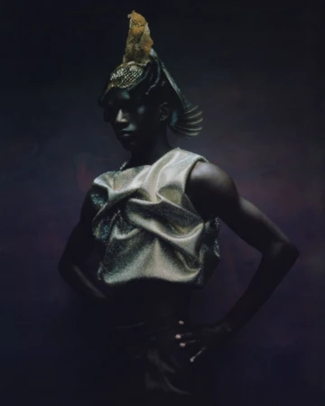 Person in a decorative golden headdress and draped metallic top poses against a dark purple backdrop in a high-fashion portrait.