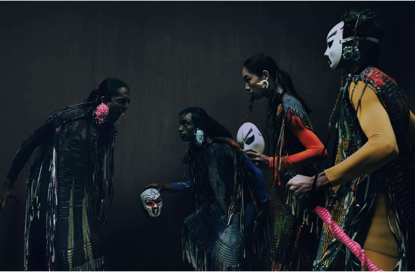 Four models in ornate, fringed costumes and theatrical masks strike stylized poses against a dark backdrop in a fashion-editorial style image.