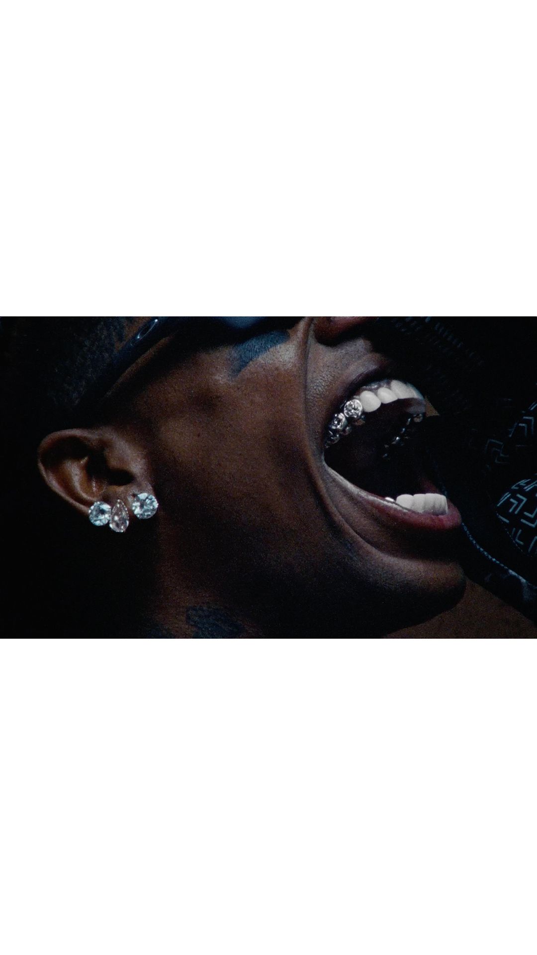 Close-up shot of a dark-skinned adult male singing, featuring a grill on his teeth and diamond earrings, captured in a dimly lit indoor setting.