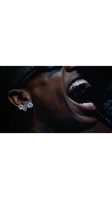 Close-up shot of a dark-skinned adult male singing, featuring a grill on his teeth and diamond earrings, captured in a dimly lit indoor setting.