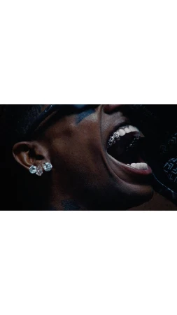 Close-up shot of a dark-skinned adult male singing, featuring a grill on his teeth and diamond earrings, captured in a dimly lit indoor setting.