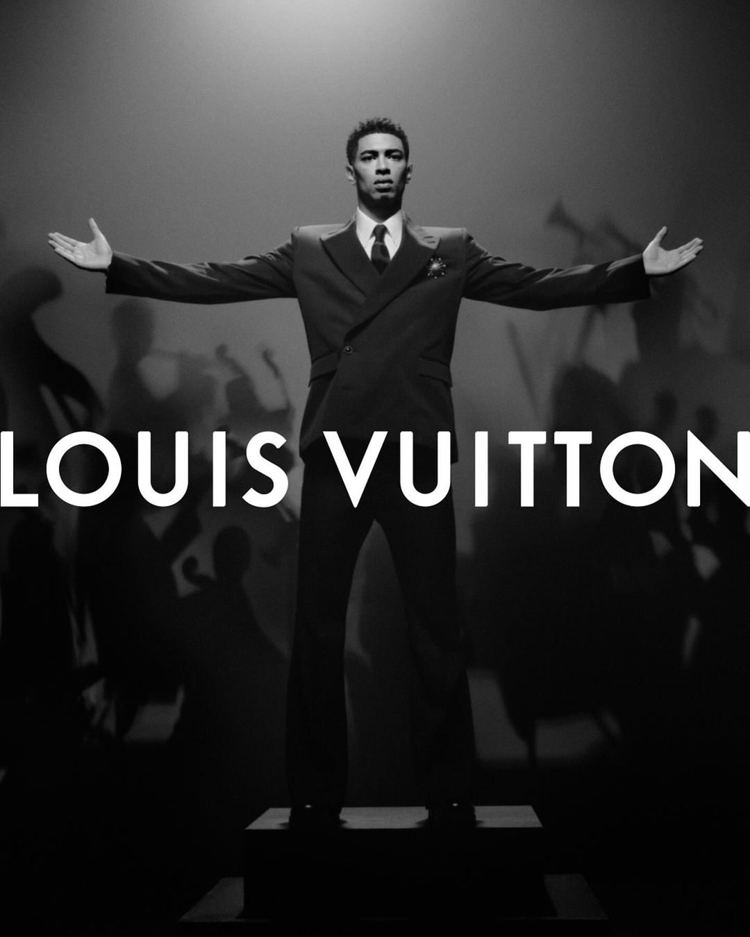 A man in a suit stands with arms outstretched against a stage background in a black and white fashion image for Louis Vuitton.
