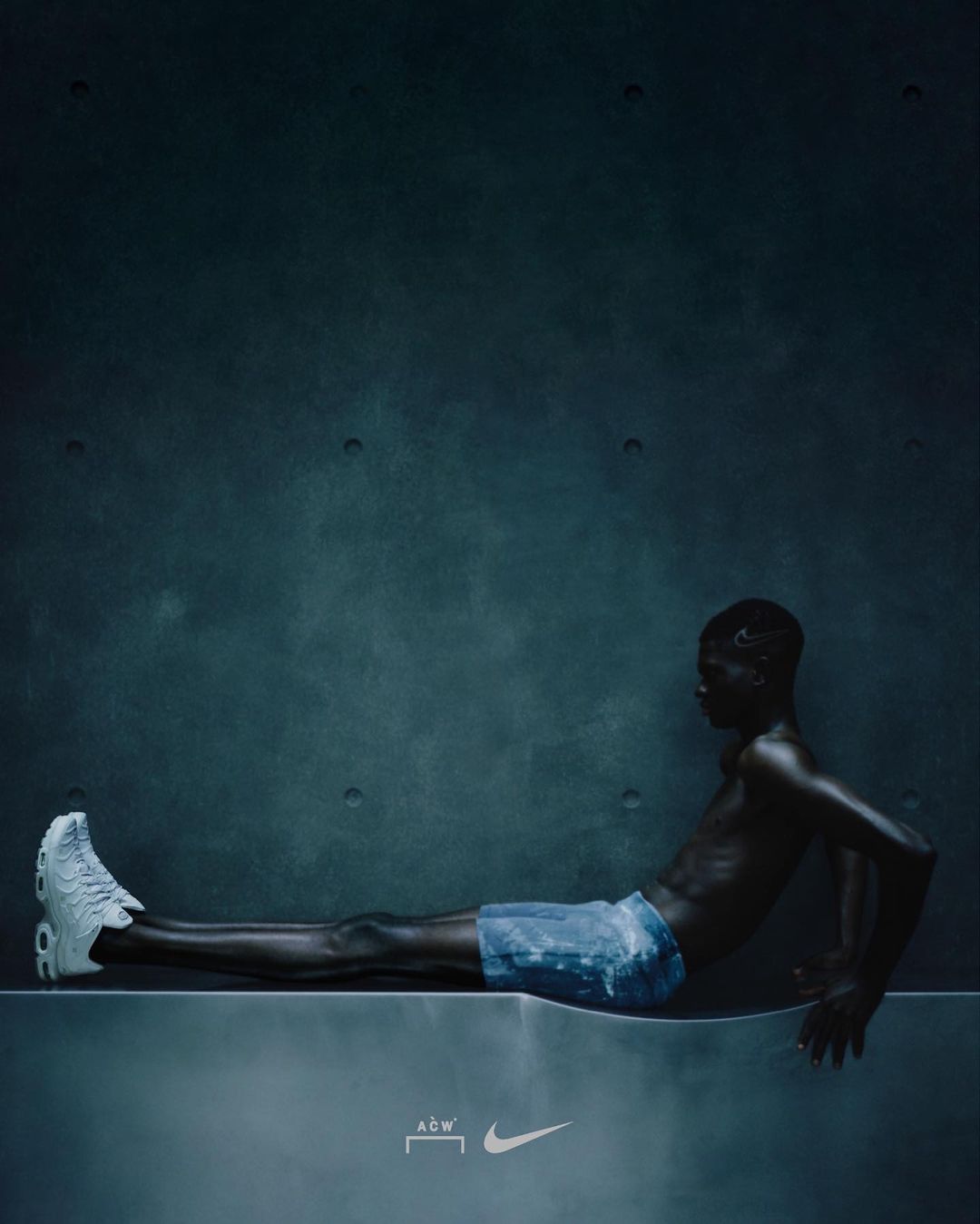 Dark image of slim African American male reclining on a concrete platform, wearing a sneaker and shorts, against a muted blue-grey wall.