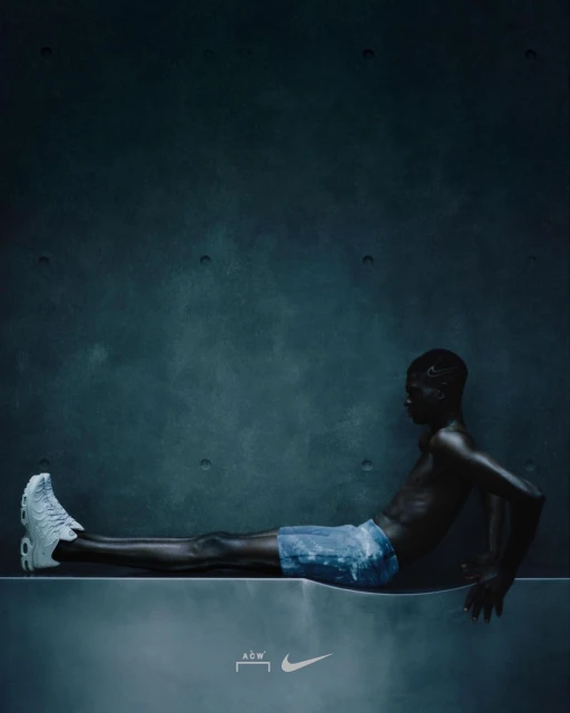 Dark image of slim African American male reclining on a concrete platform, wearing a sneaker and shorts, against a muted blue-grey wall.