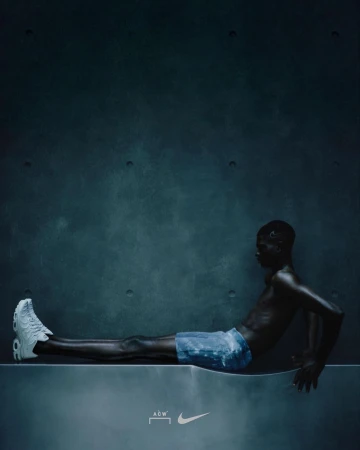Dark image of slim African American male reclining on a concrete platform, wearing a sneaker and shorts, against a muted blue-grey wall.