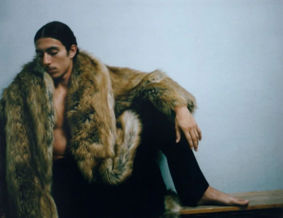 Man in fur coat sits on wooden bench against pale blue wall in studio, creating a fashion or fine art portrait with neutral mood.