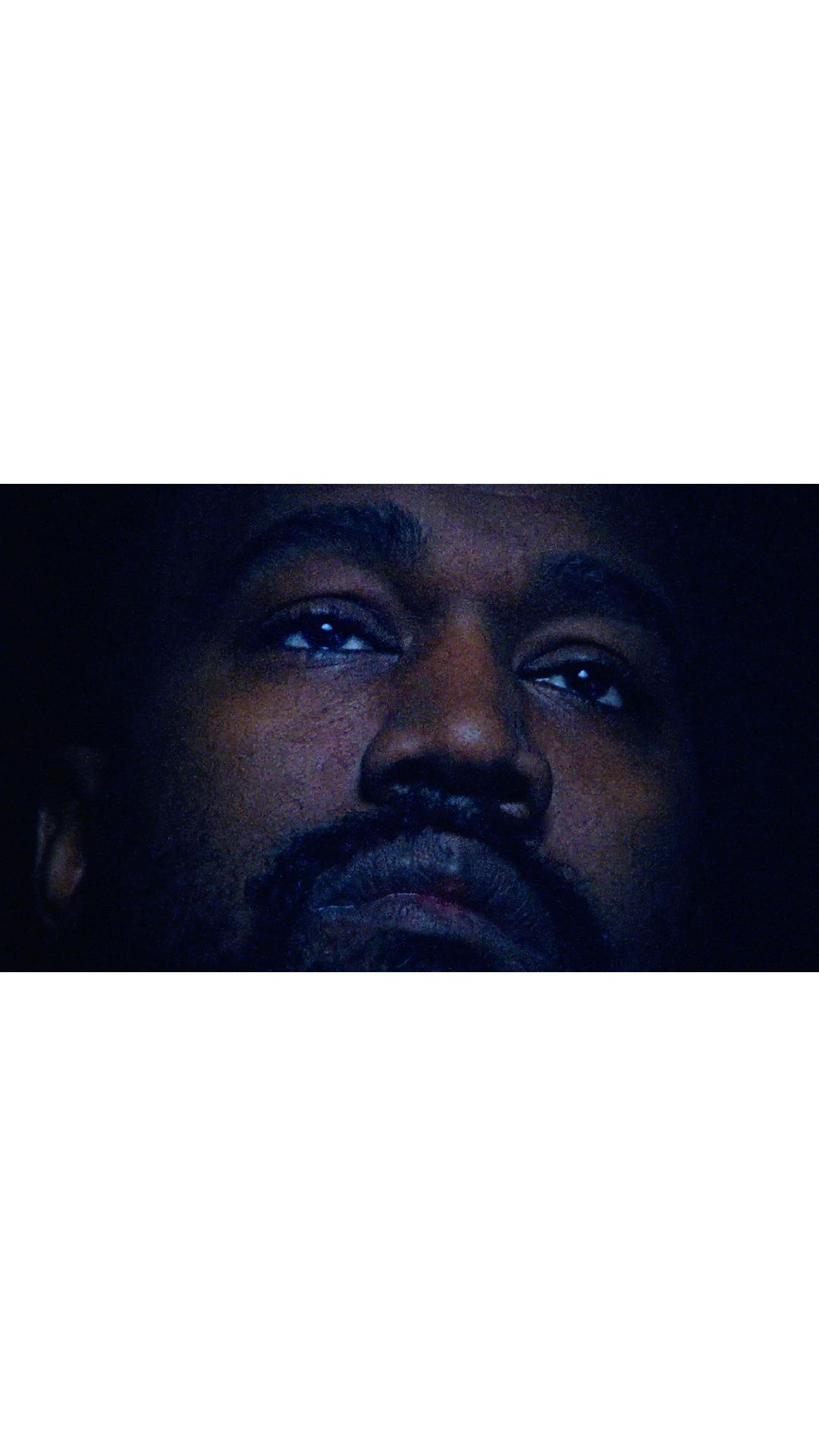 Close-up portrait of a dark-skinned adult male with a beard and mustache looking upward in a dark environment with dramatic lighting.