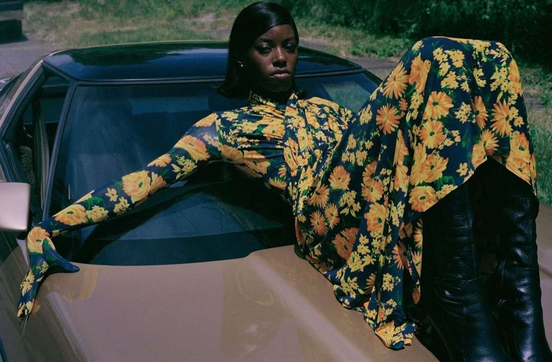 An African American woman poses on a vintage car in a floral dress and black boots, styled in a fashion editorial shoot outdoors.
