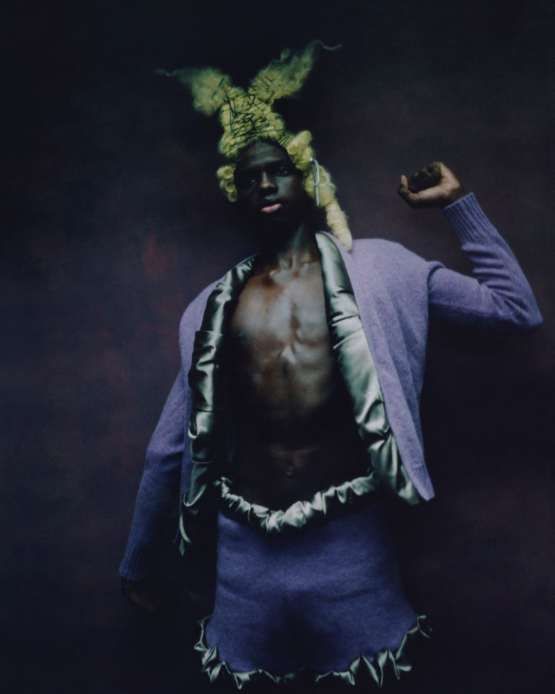 A person with dark skin poses in a purple two-piece outfit and avant-garde headpiece against a moody, dark purple background, full-body shot.