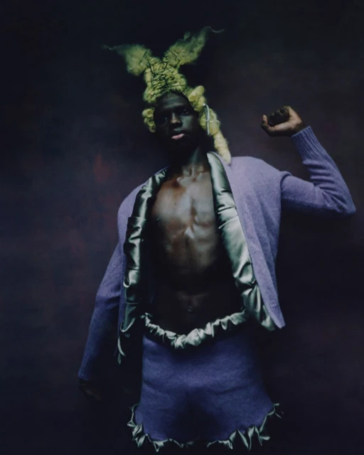 A person with dark skin poses in a purple two-piece outfit and avant-garde headpiece against a moody, dark purple background, full-body shot.