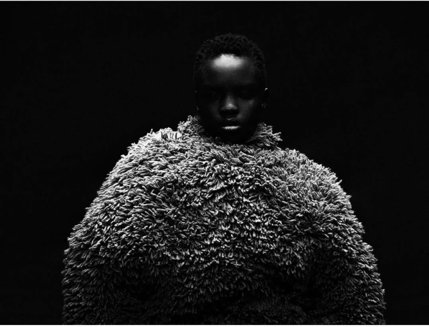 Dramatic black and white portrait of an African woman wearing a textured coat against a dark background, shot with moody lighting.
