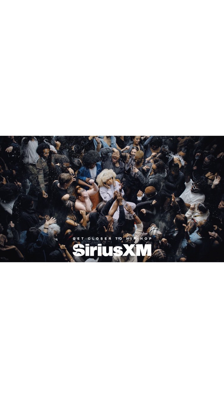 Concert crowd in low light with the SiriusXM logo in the foreground. The angle is high and shows the top of the heads of the crowd.