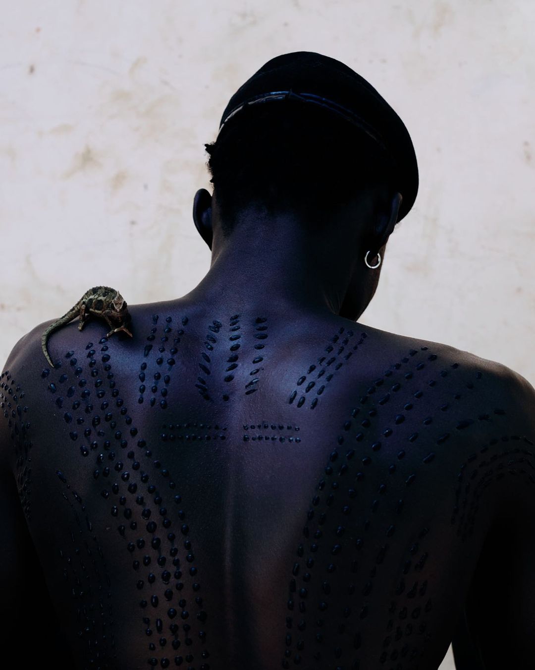 Adult man with tribal scarification and a beret poses with a chameleon on his shoulder against a neutral studio backdrop.
