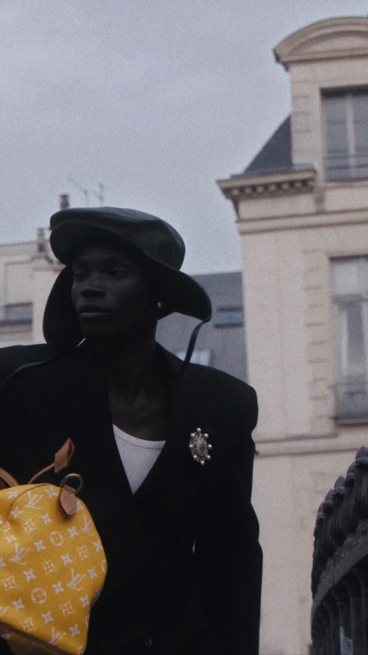 African American man in a black coat and cap walks outside with a yellow Louis Vuitton bag, with a building in the background.